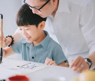 asian chinese father holding sons hand guiding him practicing chinese calligraphy at home